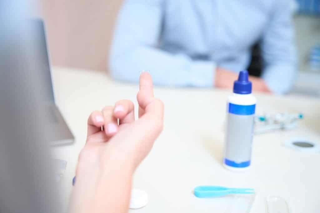 Osoba trzyma soczewkę kontaktową, obok leży płyn do pielęgnacji soczewek.